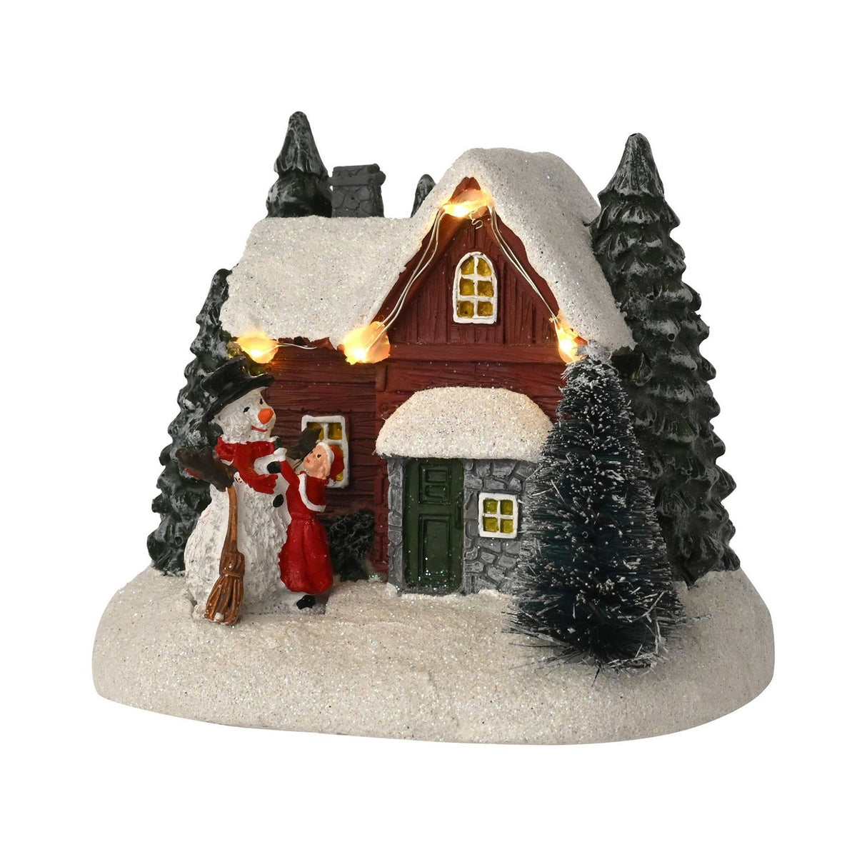 Light-up plastic house and snowman decoration with a red-walled, snow-roofed cottage, string lights, fir trees, and a child beside the snowman on a snowy base, white background.