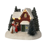 Light-up plastic house and snowman decoration with a red-walled, snow-roofed cottage, string lights, fir trees, and a child beside the snowman on a snowy base, white background.