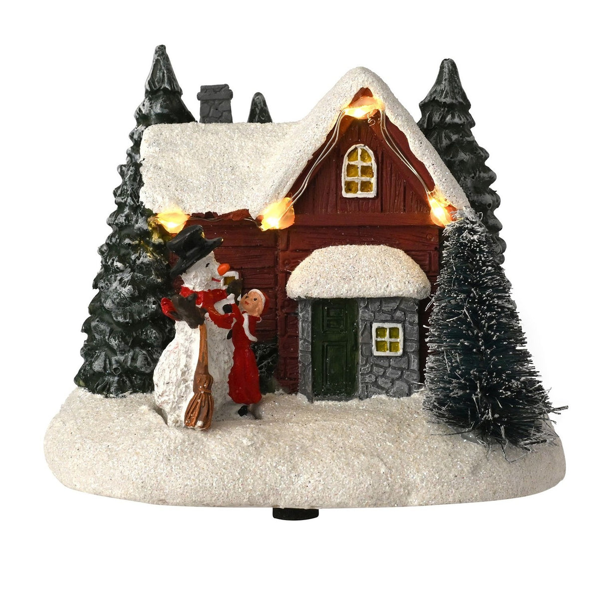 Light-up house and snowman Christmas decoration with red-walled, snow-roofed cottage, green fir trees, glowing eave lights, and a figure in red beside the snowman, on a plain white background.