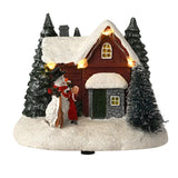 Light-up house and snowman Christmas decoration with red-walled, snow-roofed cottage, green fir trees, glowing eave lights, and a figure in red beside the snowman, on a plain white background.