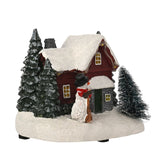 Light-up plastic Christmas decoration featuring a red-walled, snow-roofed house with bottle-brush fir trees and a top-hatted snowman holding a broom, on a white base against a plain background.