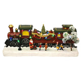 Light-up Christmas train scene decoration with red steam engine, carriages carrying presents, teddy bear and a lit tree, Santa and children figures on a snowy white base.