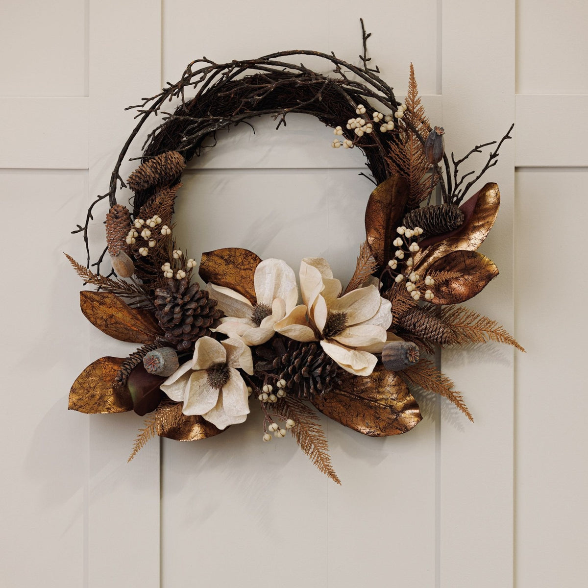 Faux Magnolia Iron Semi Wreath, 40cm, with cream magnolia blooms, pinecones, bronze leaves and bare twig branches, hung on a light neutral panelled wall.