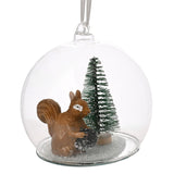 Clear glass round bauble Christmas decoration with a brown squirrel holding a pine cone beside a green bottle-brush tree on a snow-flecked base, ribbon hanger, plain white background.