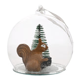 Clear glass round bauble Christmas decoration with a brown squirrel holding a pine cone beside a green bottle-brush tree on a snow-flecked base, shown on a plain white background.