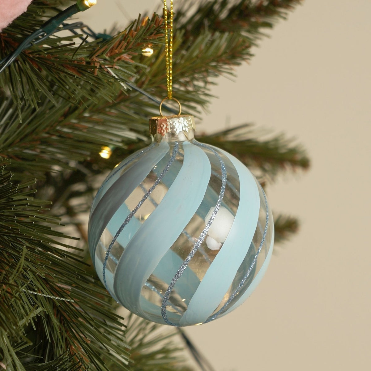 Round glass Christmas bauble with swirling frosted powder-blue stripes and fine blue glitter lines, gold cap and cord, hanging on an artificial tree with fairy lights.