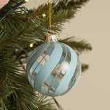 Round glass Christmas bauble with swirling frosted powder-blue stripes and fine blue glitter lines, gold cap and cord, hanging on an artificial tree with fairy lights.