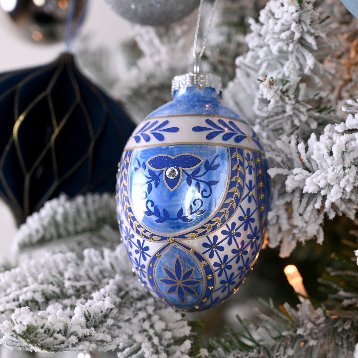 Painted glass egg Christmas tree decoration in blue with white bands, gold detailing, scrolling leaf motifs and a central clear stud, hanging on a snowy tree.