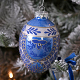 Oval painted glass egg Christmas tree decoration in blue, with white banding, gold leaf detail and a scrolling motif centred by a clear sparkling stud, shown hanging among snowy pine branches.