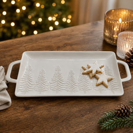 Rectangular white stoneware serving tray with handles, embossed pine tree design, holding star-shaped iced cookies on a wooden table with festive candles and lights in the background.