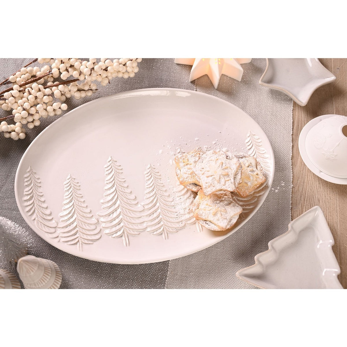 Oval stoneware serving plate in pale grey with embossed pine tree row, shown holding powdered snowflake-shaped biscuits on a festive table setting.