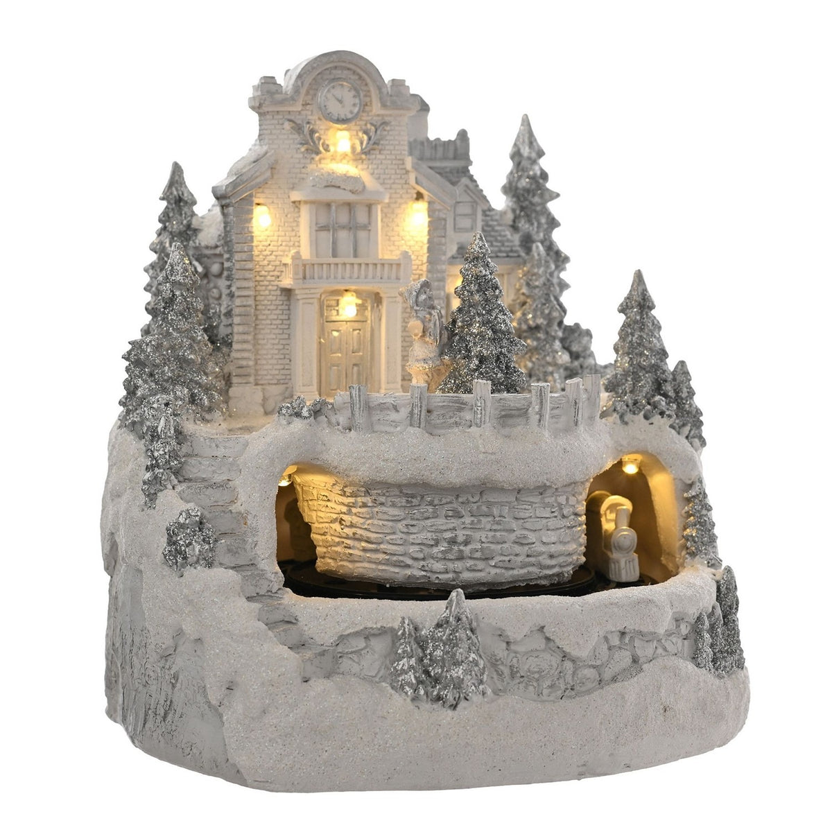 White resin Christmas village LED decoration with clocktower building, dormer-roofed house, silver glitter pine trees, fenced terrace and stone tunnel, on a plain white background.