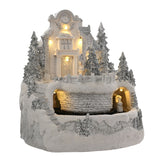 White resin Christmas village LED decoration with clocktower building, dormer-roofed house, silver glitter pine trees, fenced terrace and stone tunnel, on a plain white background.
