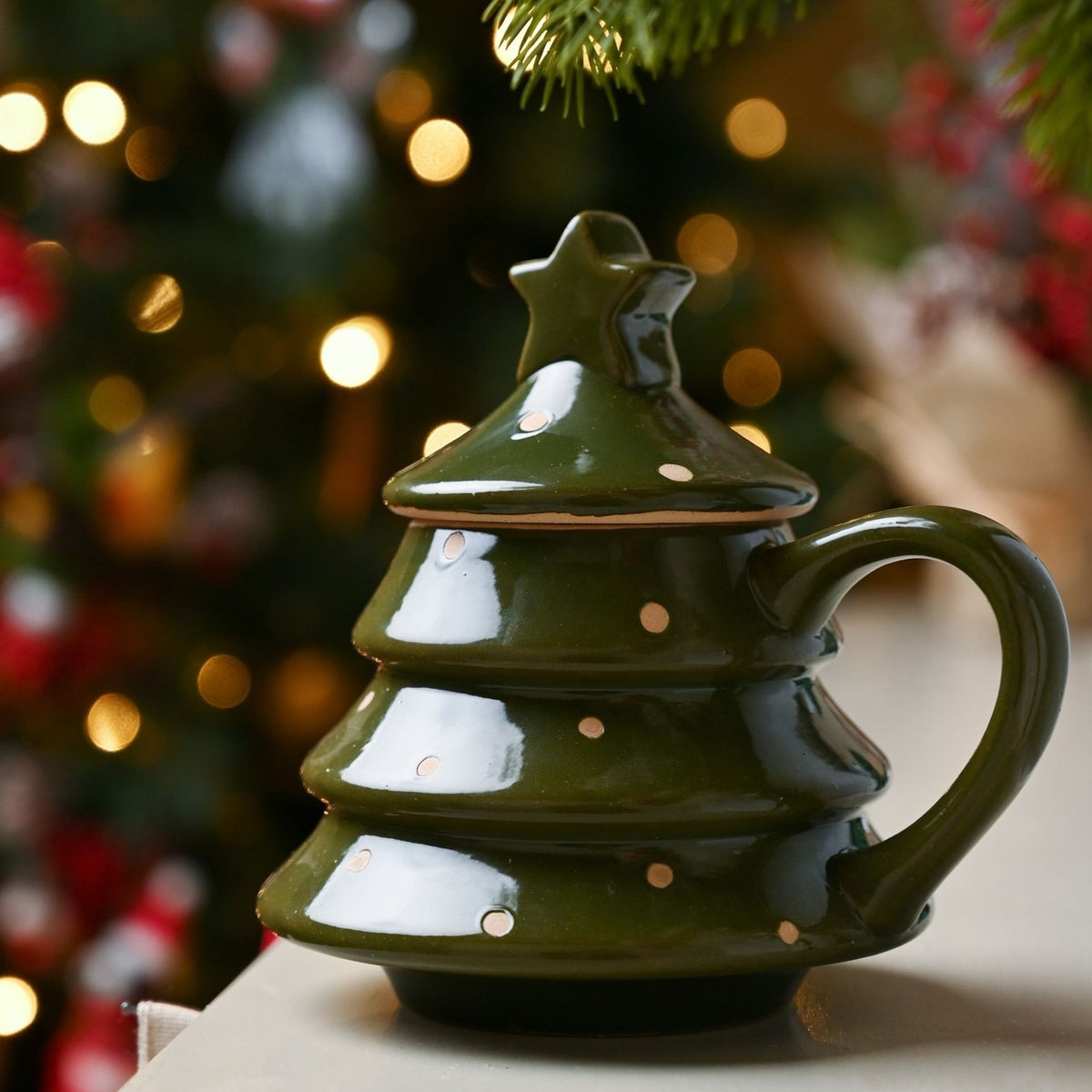 Green stoneware Christmas tree mug with polka dot design, tiered body, curved handle, and star-topped lid, shown against festive bokeh lights.