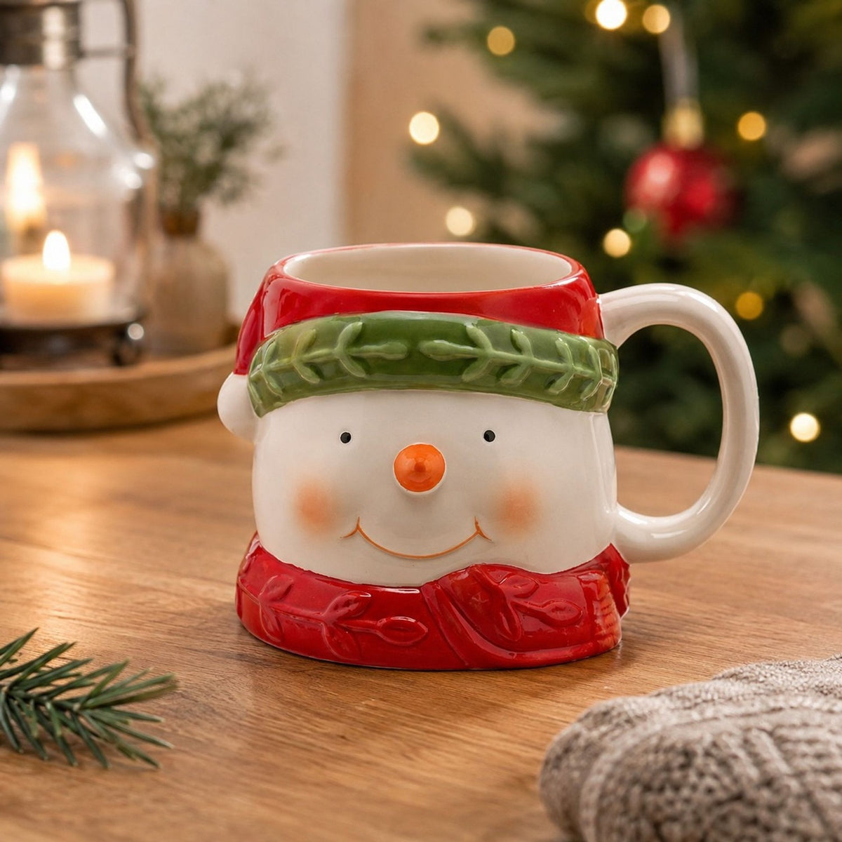 Ceramic snowman mug with white face, red hat and green band, red scarf base, and white C-handle, displayed on a wooden table with festive décor.