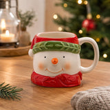 Ceramic snowman mug with white face, red hat and green band, red scarf base, and white C-handle, displayed on a wooden table with festive décor.