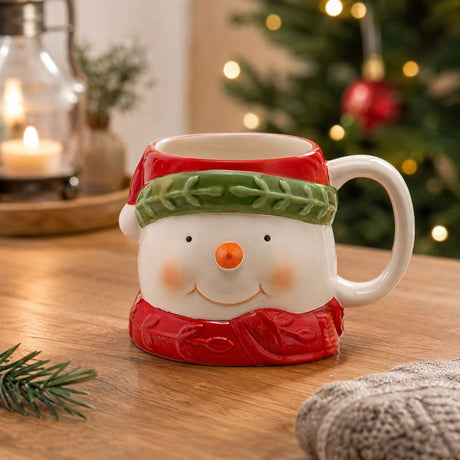 Ceramic snowman mug with white face, red hat and green band, red scarf base, and white C-handle, displayed on a wooden table with festive décor.