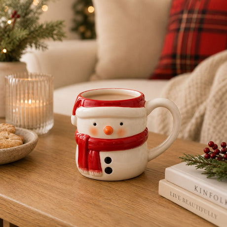 Small ceramic snowman mug with white body, red hat and scarf, black button details, filled with coffee and shown on a wooden table in a festive living room setting.