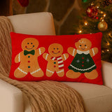 Rectangular red Christmas cushion with three gingerbread figures, featuring a bow tie, striped scarf, and a green dress with hearts and snowflakes, shown on a living room armchair.