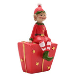 Resin elf figurine in red suit with green collar, gold buttons and belt, striped stockings and green boots, seated on a red gift box with gold stars, white background.