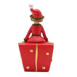 Jingle Elf on Present Christmas decoration, resin figurine in a red suit with green trim seated atop a red gift box with gold ribbon and stars, shown from behind on a white background.