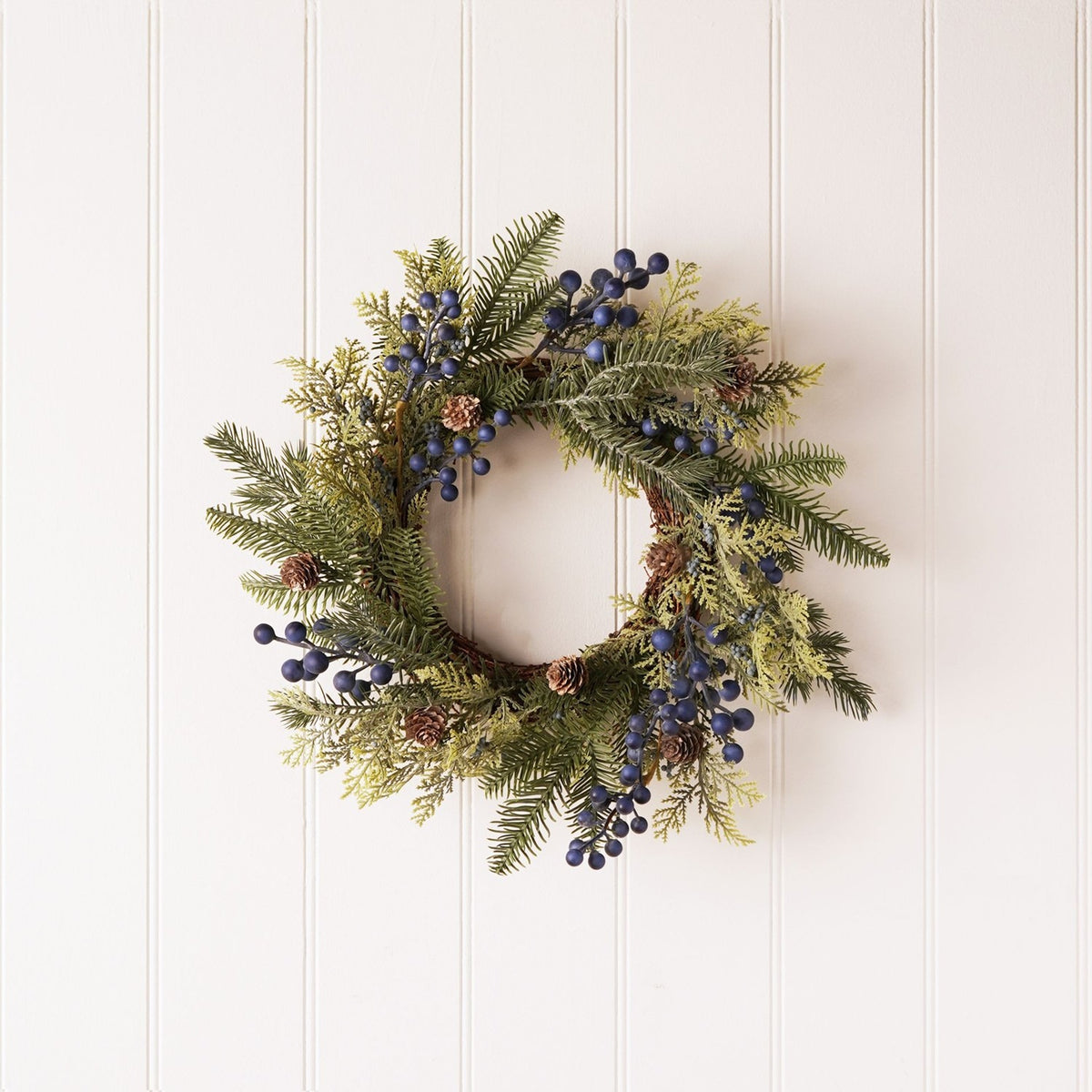 Round 40cm plastic Christmas wreath with layered green pine sprays, small brown cones, and clusters of deep blue berries, hung on a plain white panelled wall.