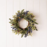 Round 40cm plastic Christmas wreath with layered green pine sprays, small brown cones, and clusters of deep blue berries, hung on a plain white panelled wall.