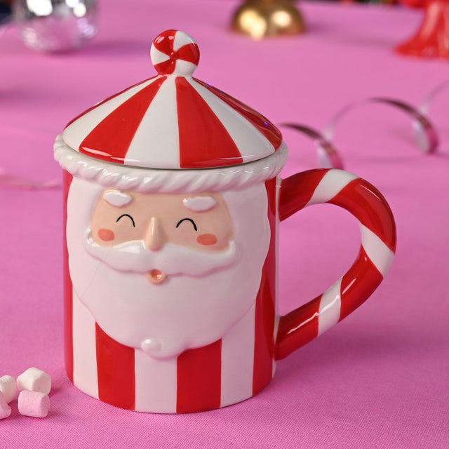 Red and white striped ceramic Santa mug with lid, featuring a raised smiling Santa face, candy-cane striped handle, and peppermint ball finial, shown on a pink surface.