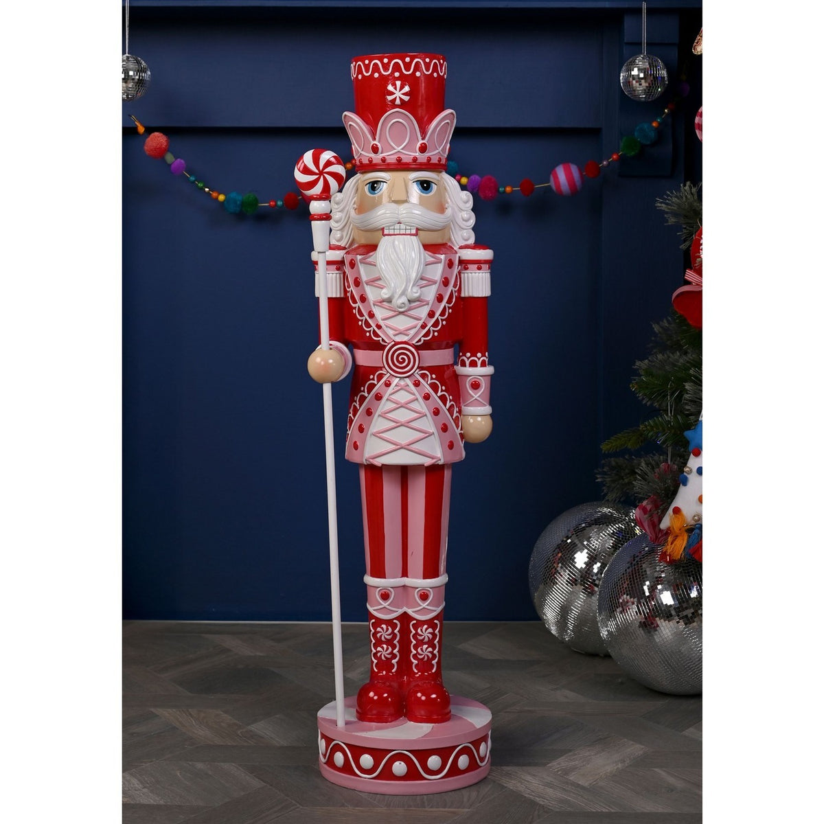 Giant 92cm resin Christmas nutcracker ornament in red and pink with white detailing, crown-band hat, white beard, candy-cane staff, standing on a round base indoors.