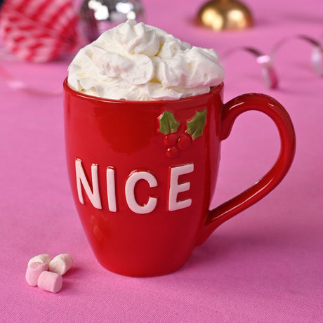 Red ceramic mug reading "NICE" in raised cream letters with a holly and berry design, filled with whipped cream, on a pink surface with marshmallows.