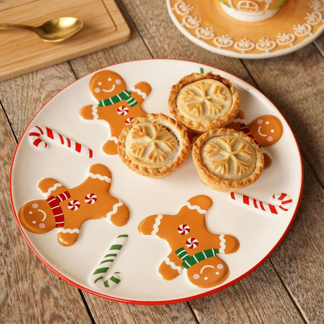 Round white ceramic dinner plate, 24cm, with red rim, decorated with gingerbread men in red and green scarves and candy canes, shown with snowflake-topped pies on a wooden table.