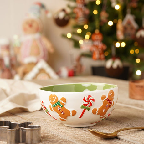 Round ceramic bowl in white with a green interior, featuring raised gingerbread men, swirl lollipops, and candy cane details, displayed on a wooden table with festive Christmas decorations.