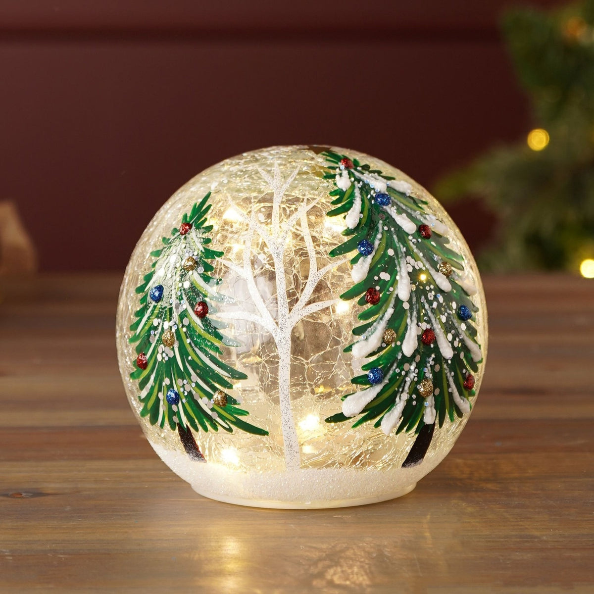 Round crackle glass light-up Christmas decoration, 12cm, painted with two snow-laden evergreen trees and a bare white tree on a frosted base, lit from within, shown on a wooden surface.