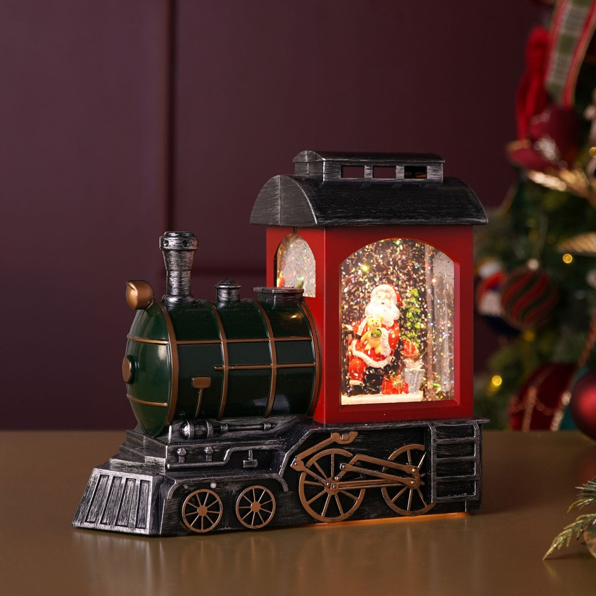 LED Glitter Santa Train Light-Up Decoration, 21.5cm, with a green boiler, red cab under an antiqued silver roof, and Santa visible inside beside a tree, shown on a table near a Christmas tree.