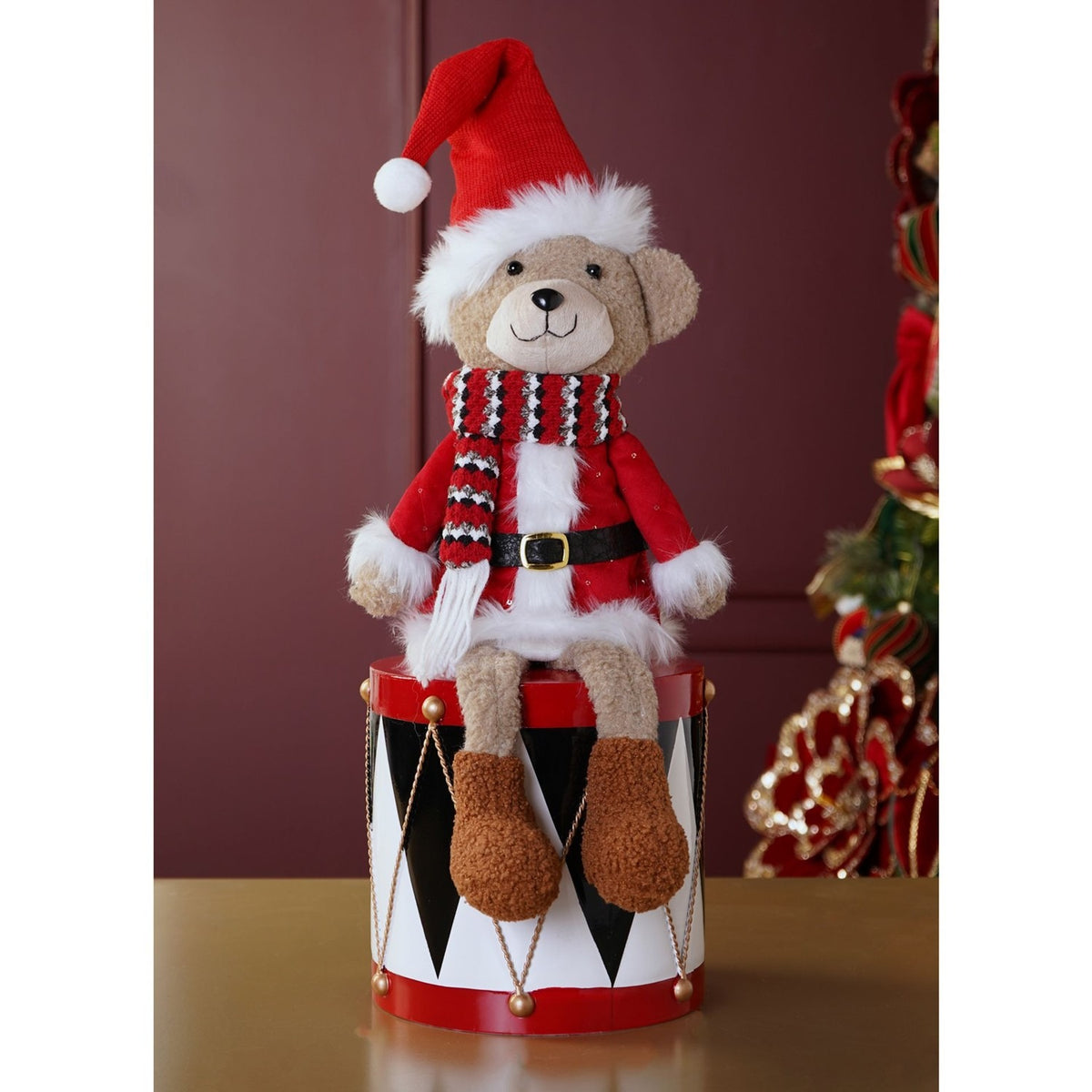 Plush sitting bear Christmas decoration in a red Santa coat with white trim, red and white striped scarf, and red Santa hat, displayed on a toy drum against a dark red wall.