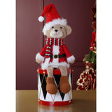 Plush sitting bear Christmas decoration in a red Santa coat with white trim, red and white striped scarf, and red Santa hat, displayed on a toy drum against a dark red wall.
