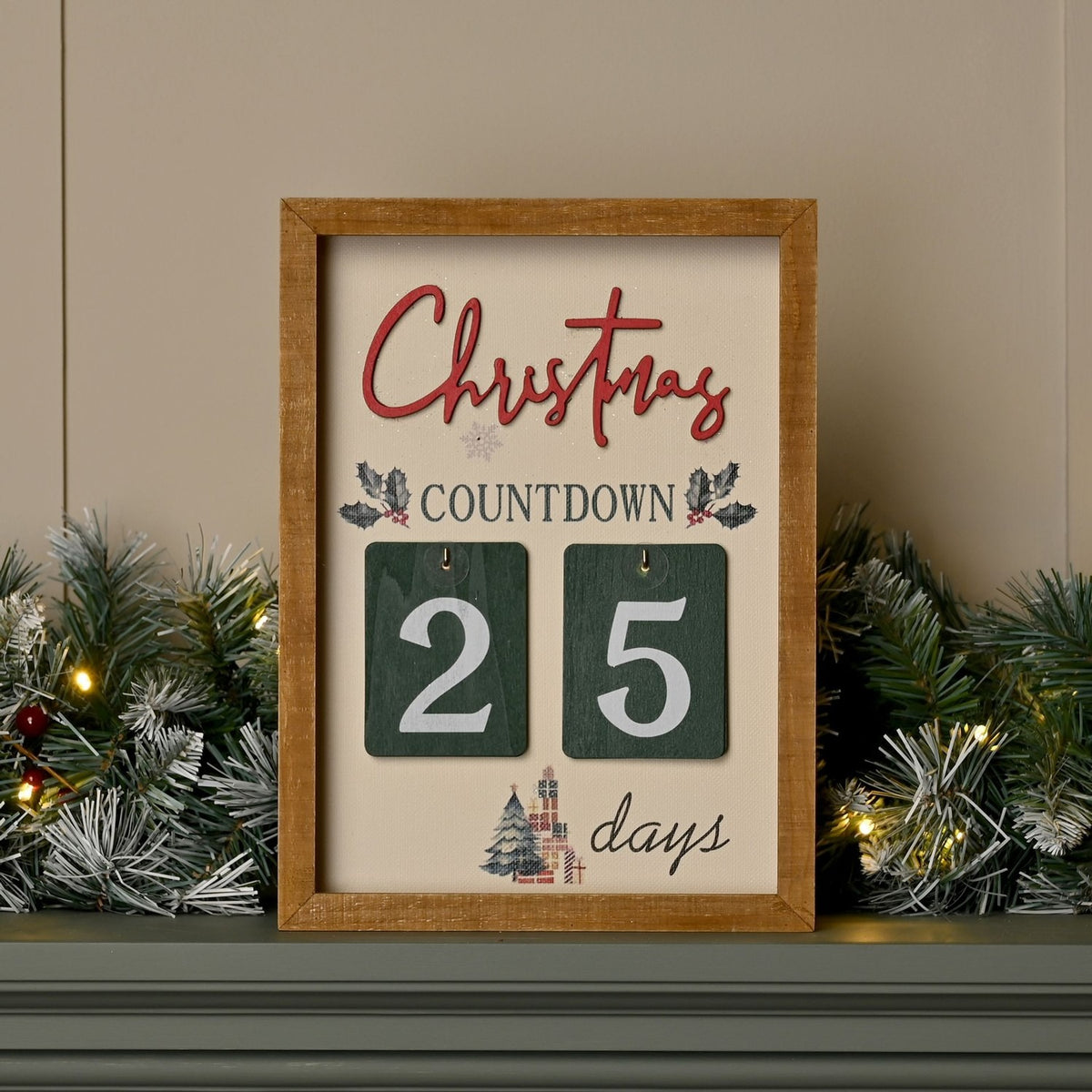 Wooden Christmas countdown plaque with cream panel, red Christmas script, holly sprigs, and hanging green number tiles showing 25 days, displayed on a garland-decorated mantel.