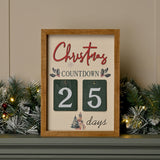 Wooden Christmas countdown plaque with cream panel, red Christmas script, holly sprigs, and hanging green number tiles showing 25 days, displayed on a garland-decorated mantel.