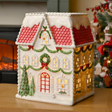 Frosted LED house light-up Christmas decoration in white plastic with a red scalloped roof, glowing windows, and green garlands, displayed on a wooden surface near a tree.