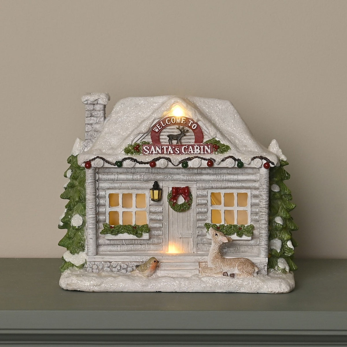 Snowy Santa Cabin light-up decoration in white and grey resin, with glittering roof, stone chimney, fir trees, wreath, lantern, deer and robin figures on a mantel.