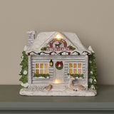 Snowy Santa Cabin light-up decoration in white and grey resin, with glittering roof, stone chimney, fir trees, wreath, lantern, deer and robin figures on a mantel.