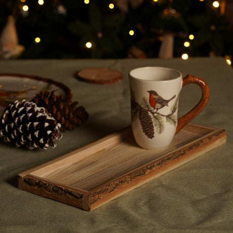 Small rectangular wooden tray with natural bark edges, holding a robin-print mug on a festive table with pine cones and fairy lights in the background.