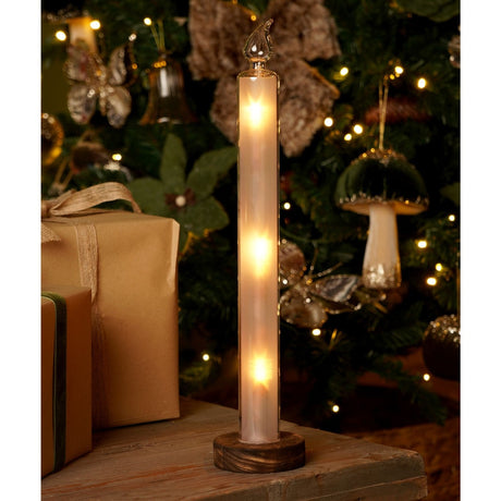 Fire Rising Effect Glass LED Taper Candle, 39cm, with frosted white column, wooden base and clear glass flame tip, glowing warm amber beside gifts and a Christmas tree.