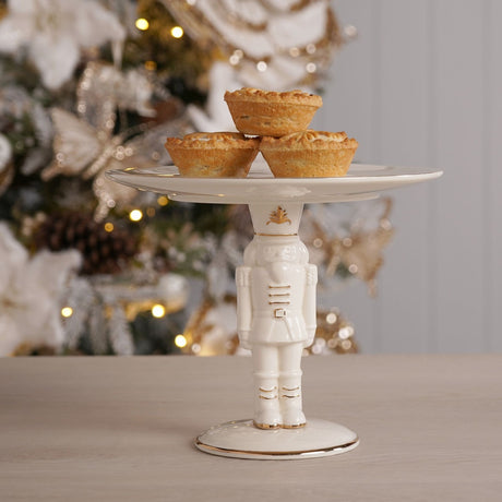 White ceramic Nutcracker cake stand with gold trim, soldier figure base holding a round plate topped with pies, Christmas tree setting.