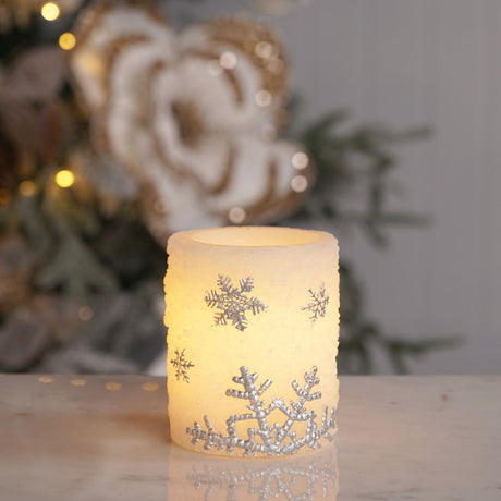 Round white LED pillar candle, 10cm, with a textured plastic finish and raised silver-grey snowflake details, glowing on a marble surface near festive décor.