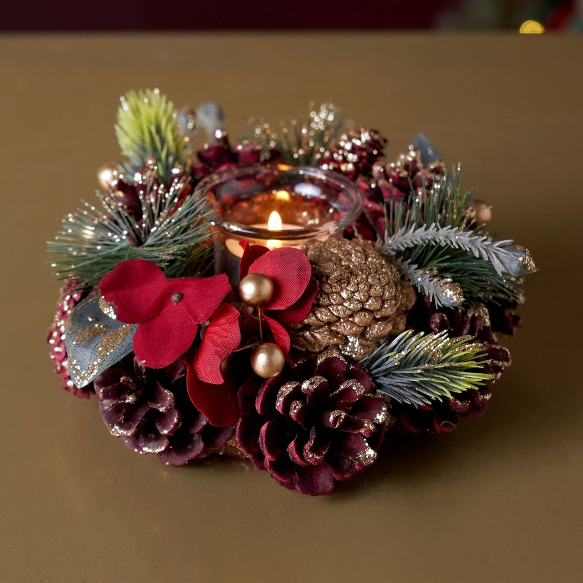 Pinecone wreath candle holder, 20cm, with a single clear glass tealight cup encircled by red and gold glittered pinecones, pine sprigs, red petals and gold berries, lit candle shown on a tabletop.