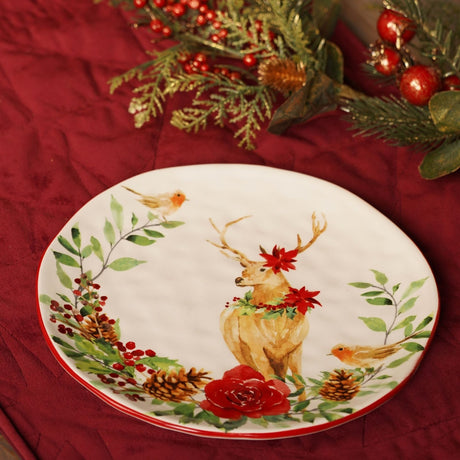 Round white ceramic dinner plate, 22cm, red rim, watercolour stag, robins, poinsettias, berries, pine cones and red rose, on red fabric with festive greenery.