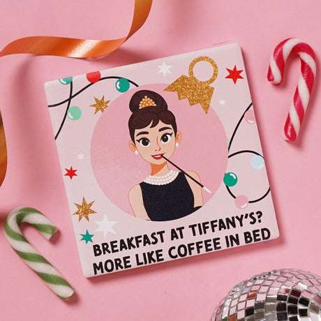 Pink ceramic coaster featuring a cartoon woman in a black dress and pearls with the text "Breakfast at Tiffany's? More Like Coffee in Bed," set against candy canes and a disco ball.