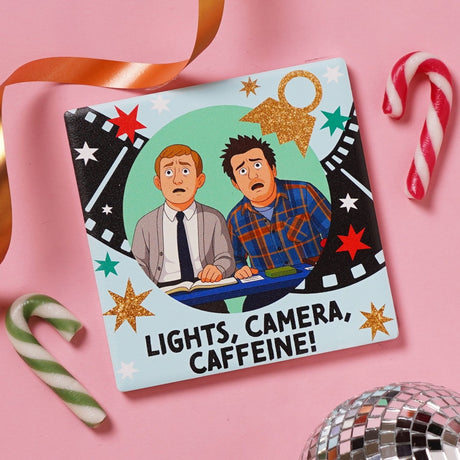 Light blue ceramic coaster with a film strip and star design, cartoon characters at a desk, Lights, Camera, Caffeine text, pink background with candy canes and a disco ball.