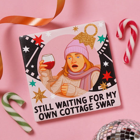 Square ceramic coaster printed with a woman in a lilac beanie holding red wine beside "Still waiting for my own cottage swap," framed by film reel strips on pink, shown with candy canes and ribbon.
